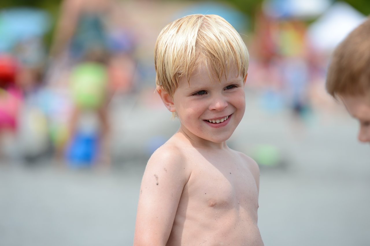 A young boy with wet blond hair smiles while standing shirtless on a busy beach.