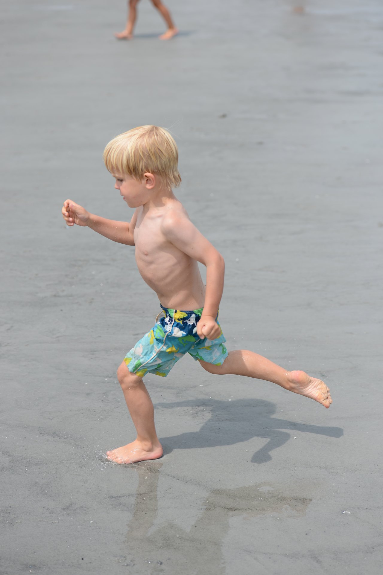 A young child in swim trunks runs barefoot on wet sand at the beach.