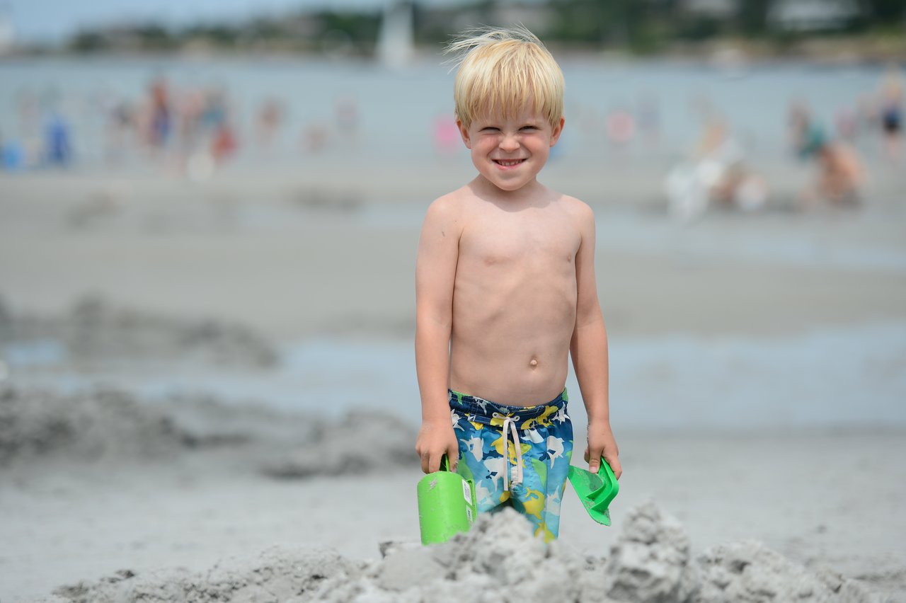 A young child in swim trunks stands on the beach, holding green sand toys and smiling at the camera.