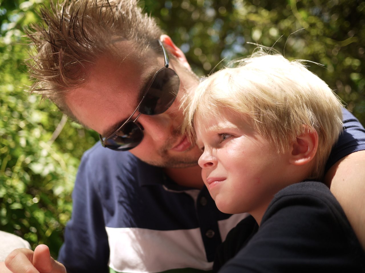 A man wearing sunglasses embraces a young boy outdoors, both looking in the same direction with serious expressions.