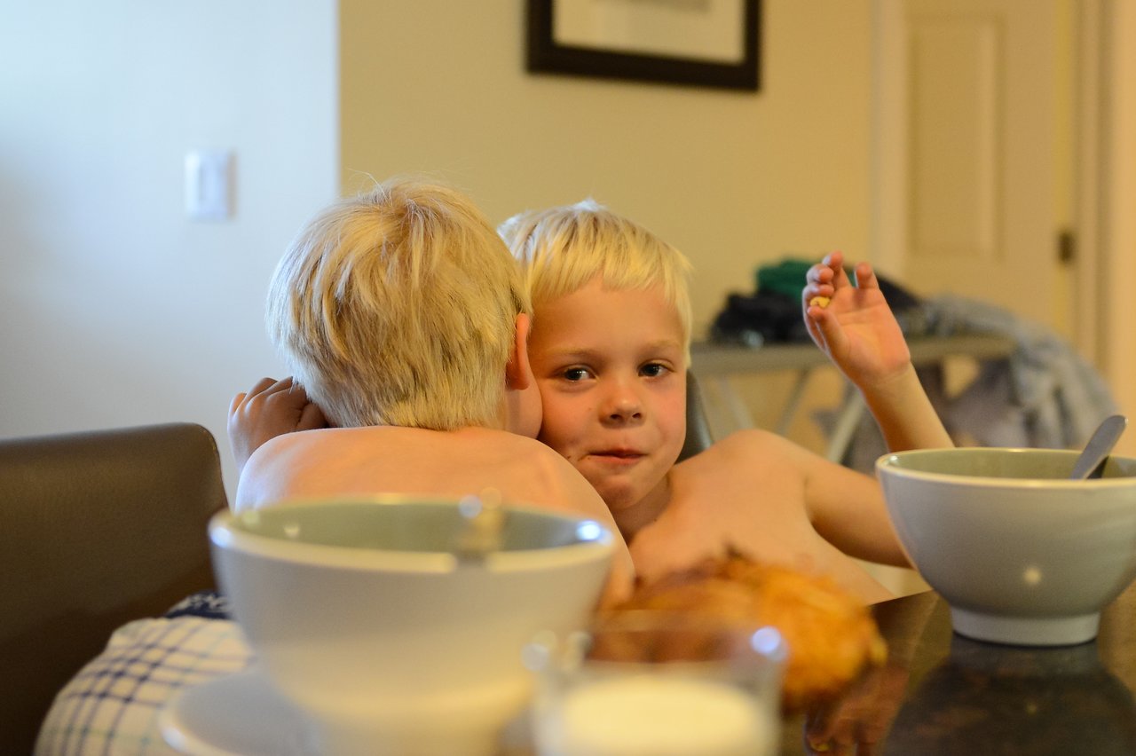 Two young, shirtless children with blonde hair sit at a table; one hugs the other while eating breakfast.
