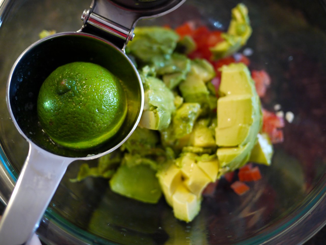 A lime is being squeezed over chopped avocado and diced tomatoes in a glass bowl.