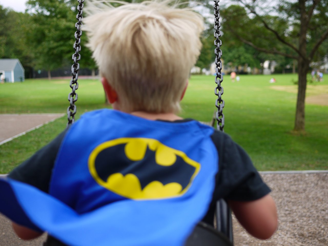 A child wearing a Batman cape swings at a park, facing away from the camera.