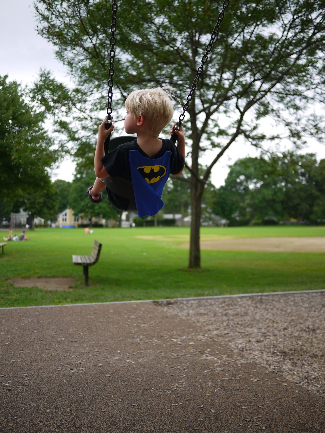 A child wearing a Batman cape swings at a park, holding the chains and looking forward.