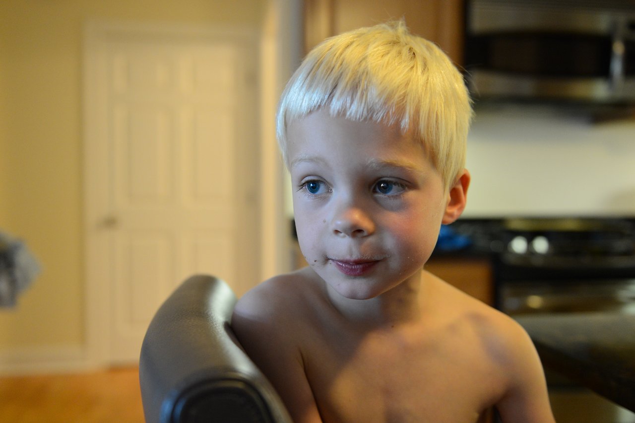 A young child with short blond hair sits shirtless on a chair, looking off to the side.
