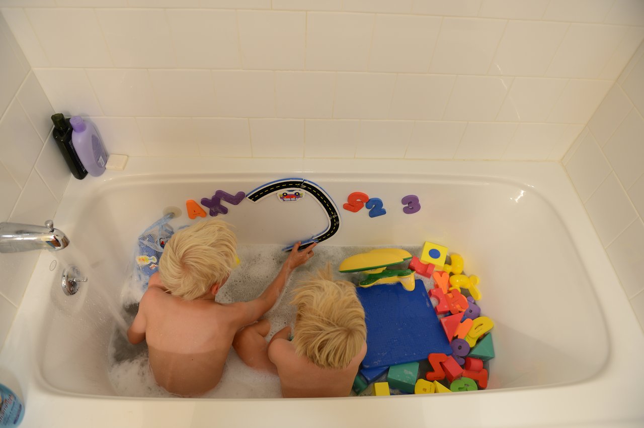 Two young children with blonde hair play with colorful foam letters and toys in a bathtub filled with water.
