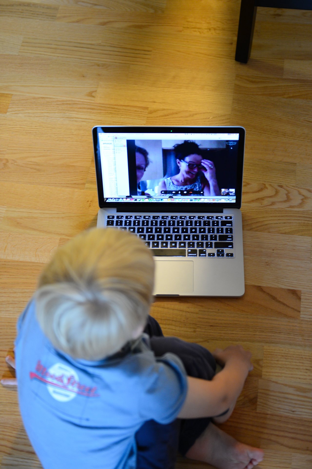 A child sits on the floor watching a video call on a laptop showing two people on the screen.