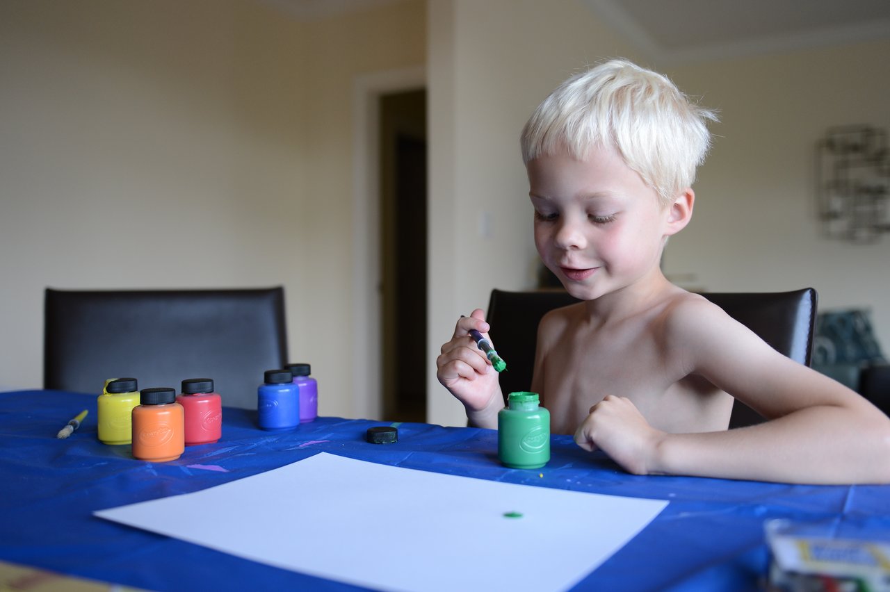 A young child sits at a table, dipping a brush into green paint, preparing to create artwork.