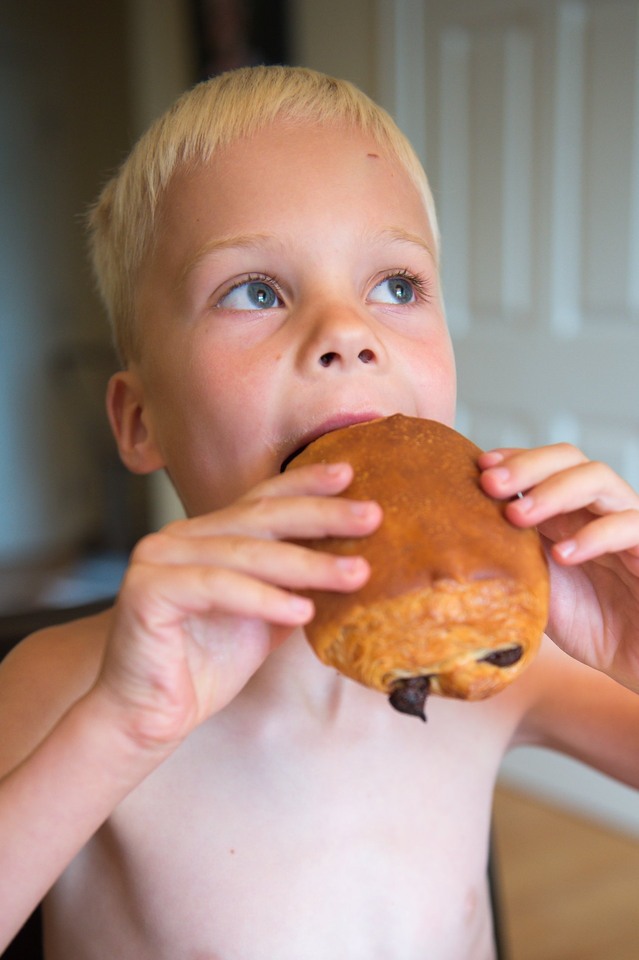 A child holds a chocolate croissant with both hands and takes a bite.