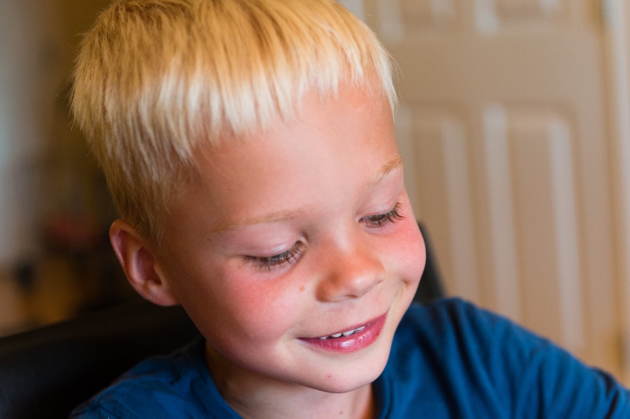 A young child with short blond hair and a blue shirt smiles and laughs while looking down.