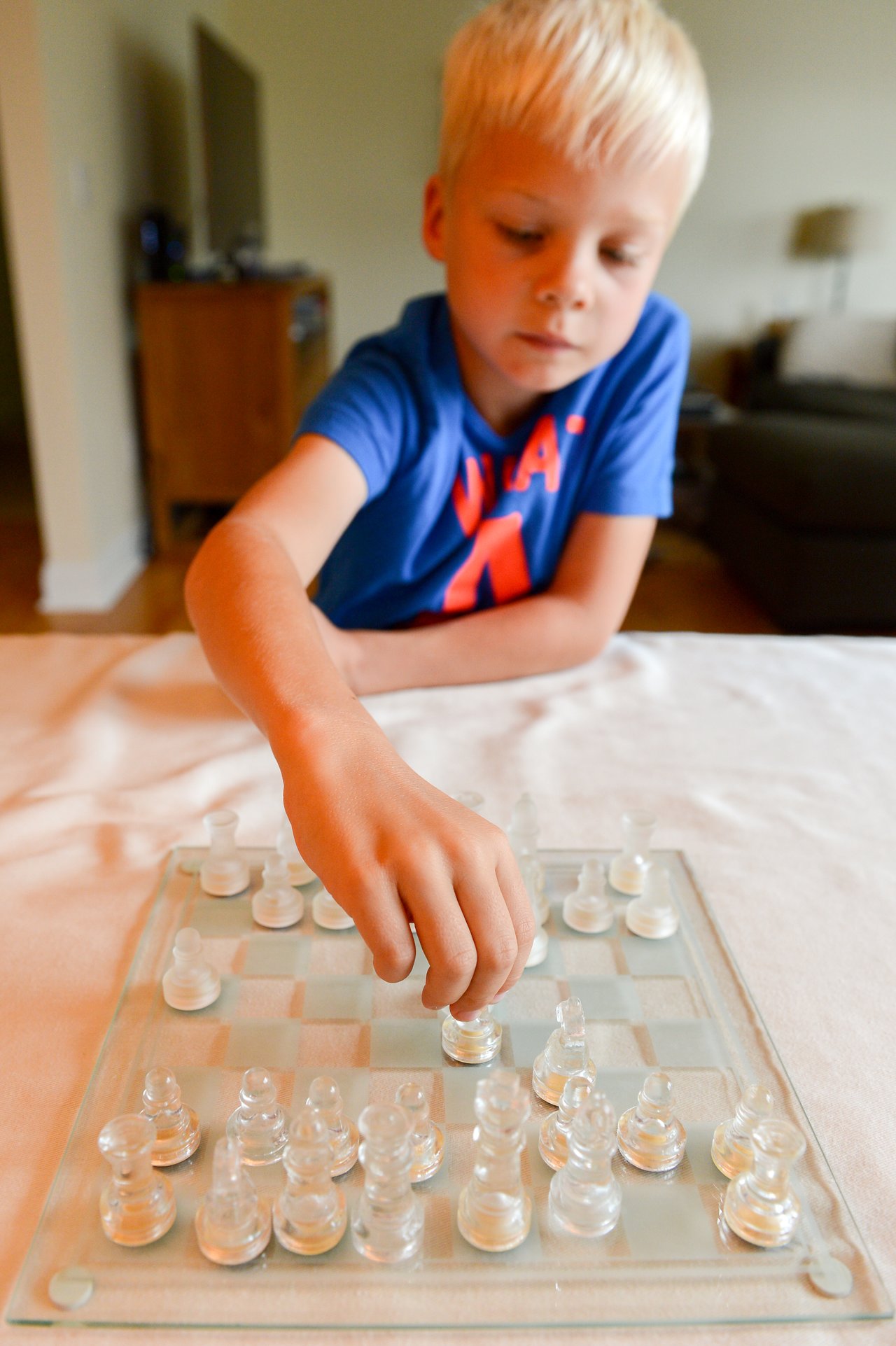 A young boy in a blue shirt moves a piece on a glass chessboard, focusing on the game.