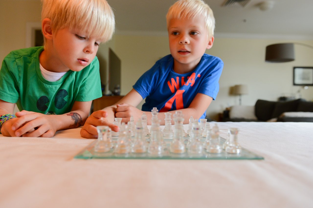 A young boy in a blue shirt plays chess with another boy in a green shirt, both focused.