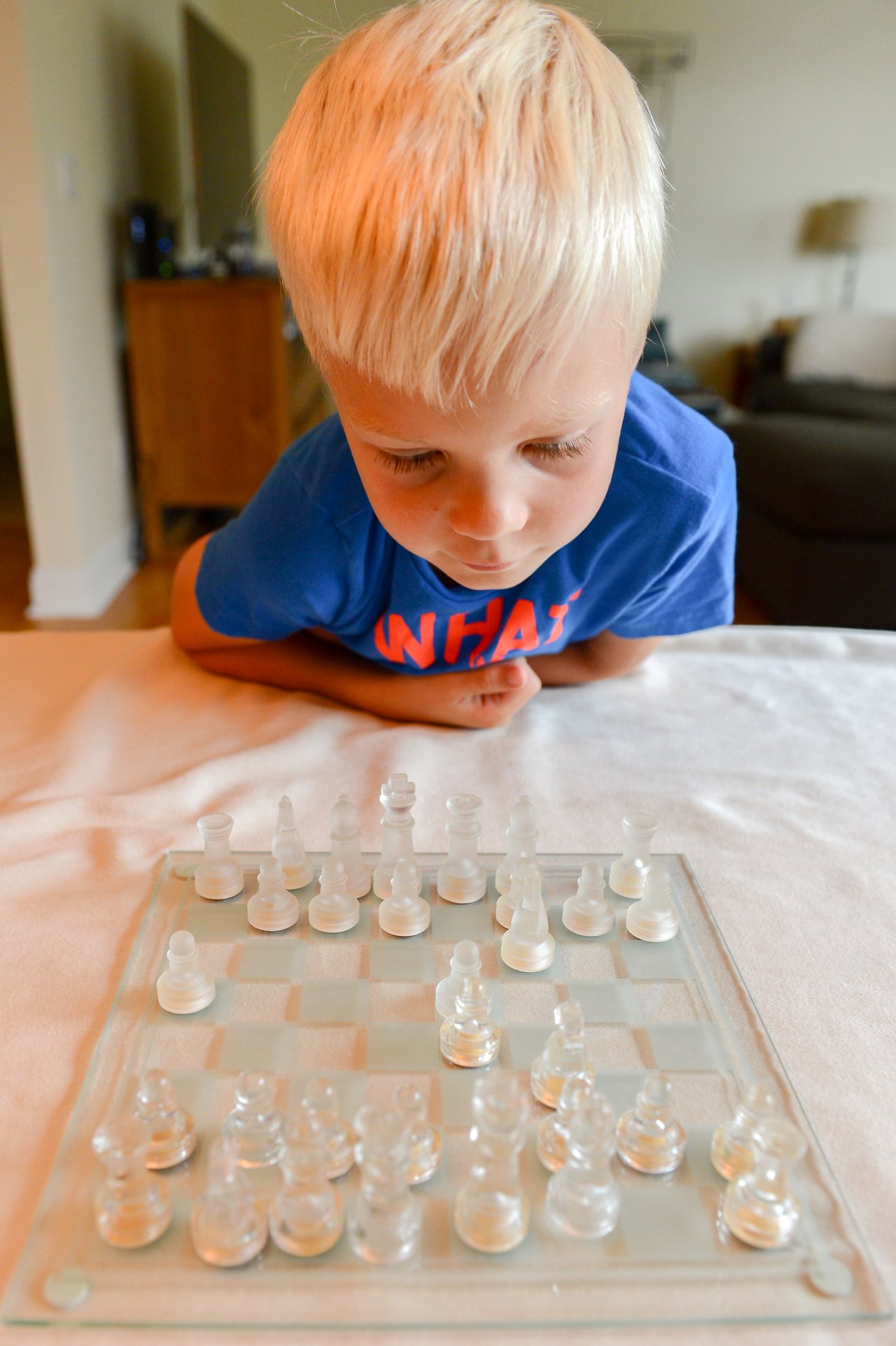 A young child in a blue shirt leans over a glass chessboard, focusing on the game pieces.