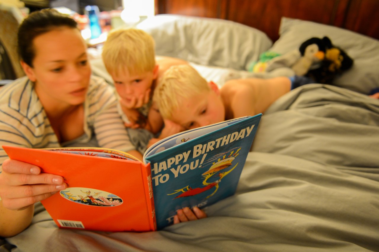 A woman reads a bedtime story to two young children lying in bed, both focused on the book.