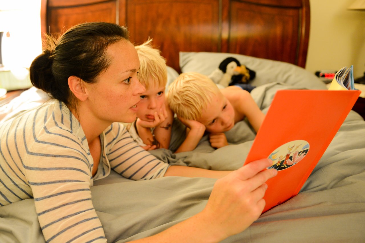 A woman reads a bedtime story to two young children lying in bed, listening closely.