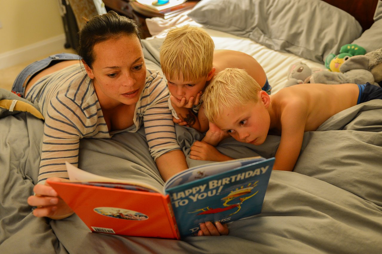 A woman reads a bedtime story to two young boys lying on a bed, both listening closely.