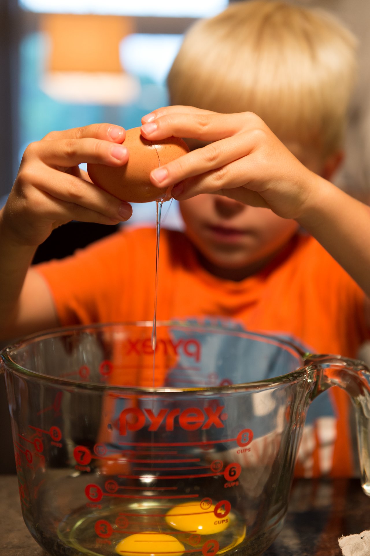 A child in an orange shirt cracks an egg into a glass measuring cup.