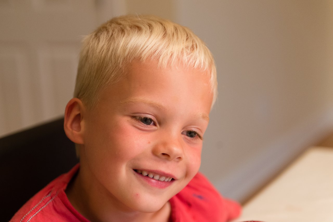 A smiling child in a red shirt sits at a table, looking happy during a birthday celebration.