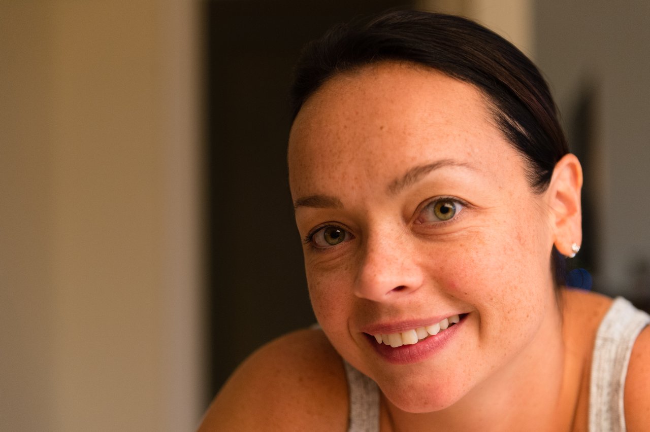 A person with dark hair and a smile looks directly at the camera in a warmly lit indoor setting.