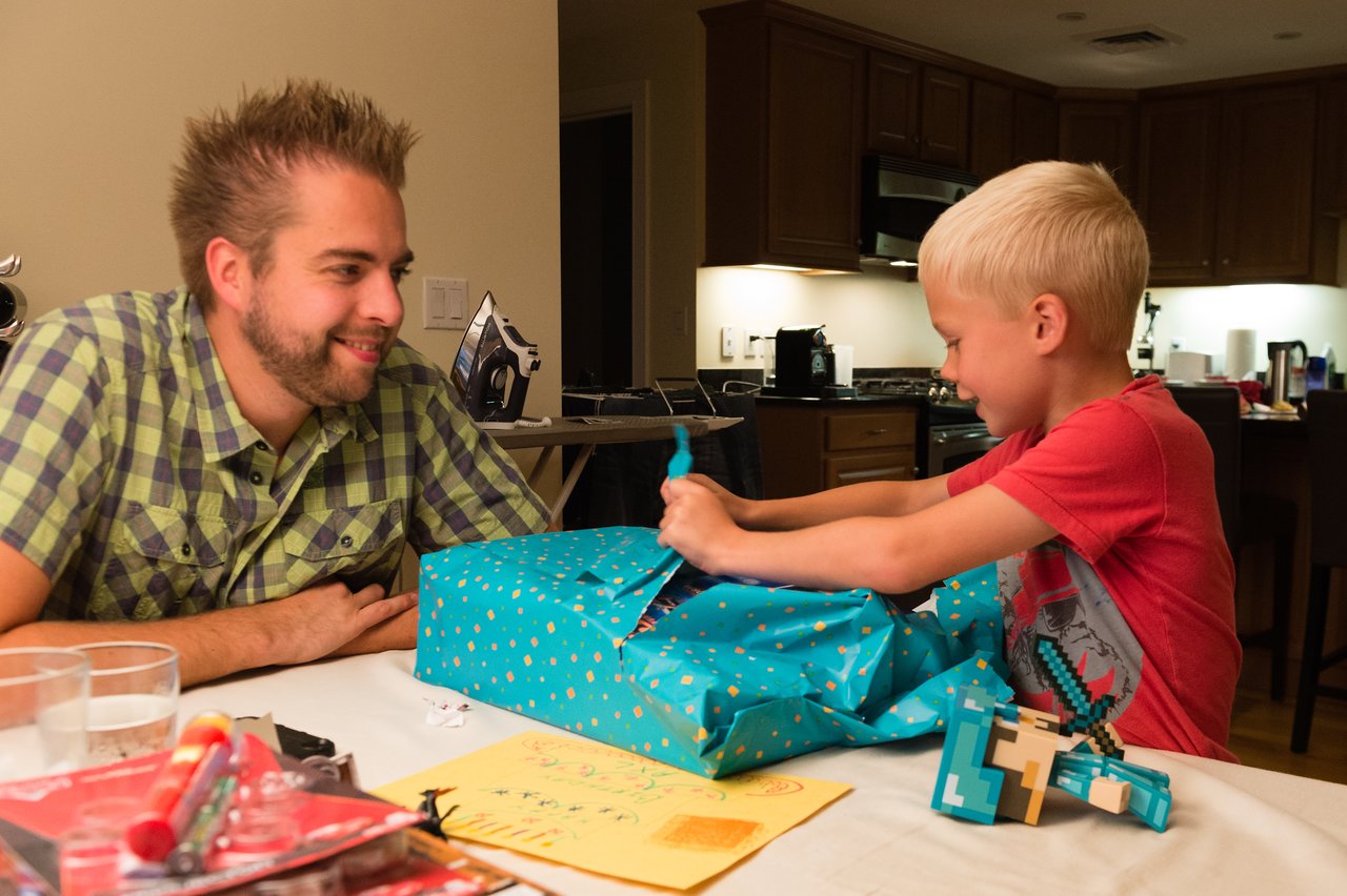 A young boy excitedly opens a birthday gift while an adult watches and smiles at him.