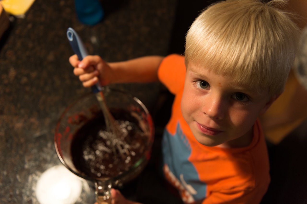 A young child in an orange shirt stirs chocolate batter in a glass bowl while looking up at the camera.