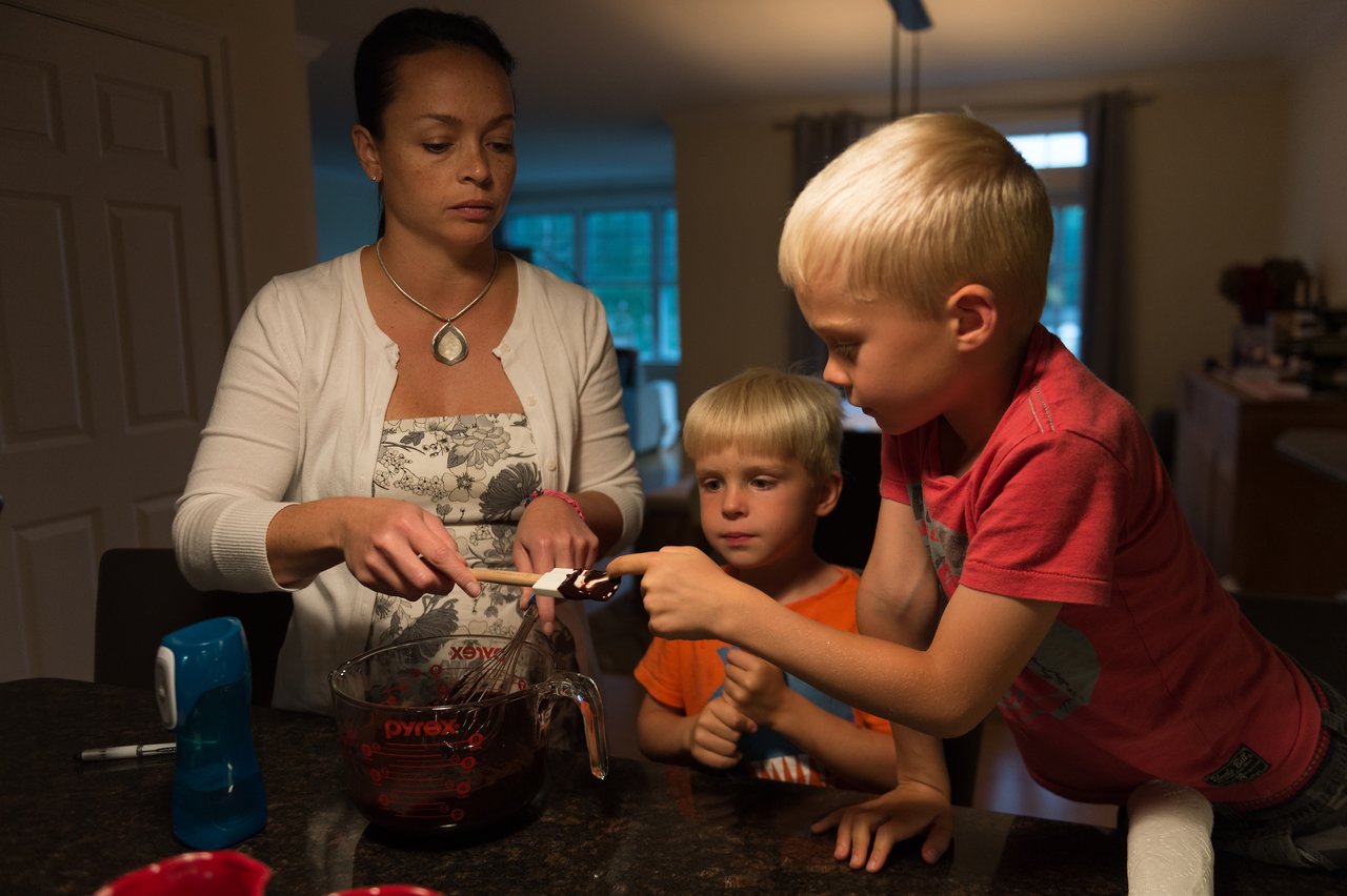 A woman and two children mix ingredients in a measuring cup, preparing something together in the kitchen.