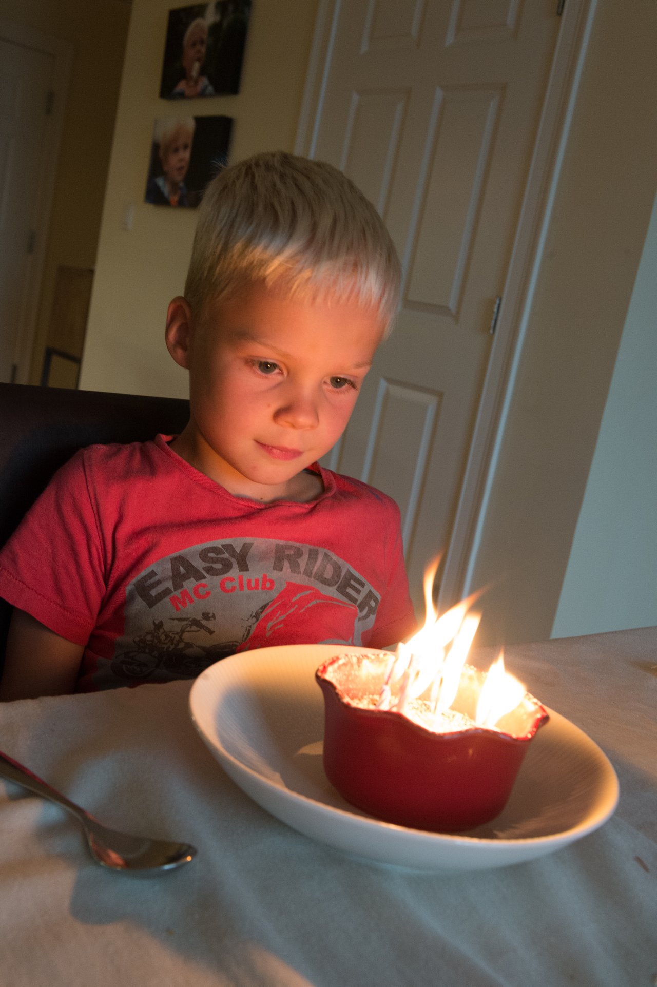 A young child in a red shirt looks at a small birthday dessert with lit candles on the table.