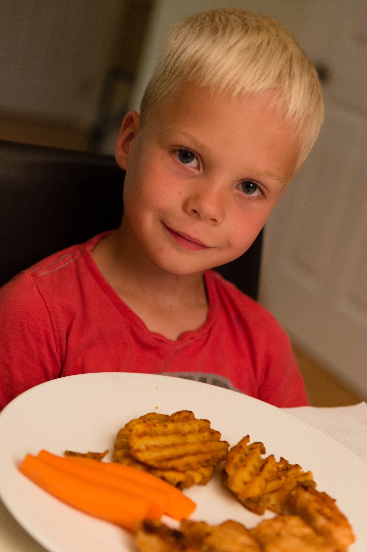 A child in a red shirt sits at a table, smiling with a plate of food in front of them.