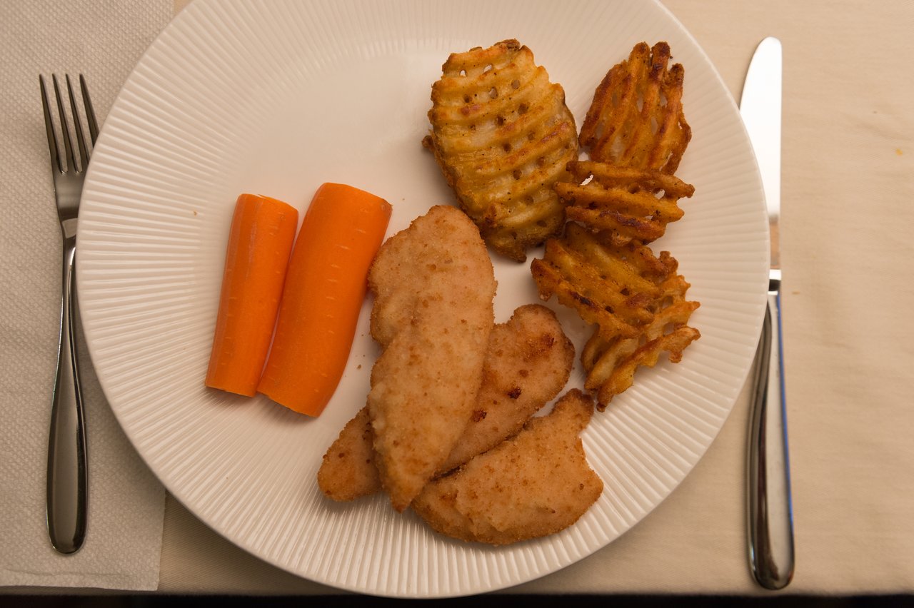 A white plate with breaded chicken, waffle fries, and carrot sticks on a table with utensils and a napkin.