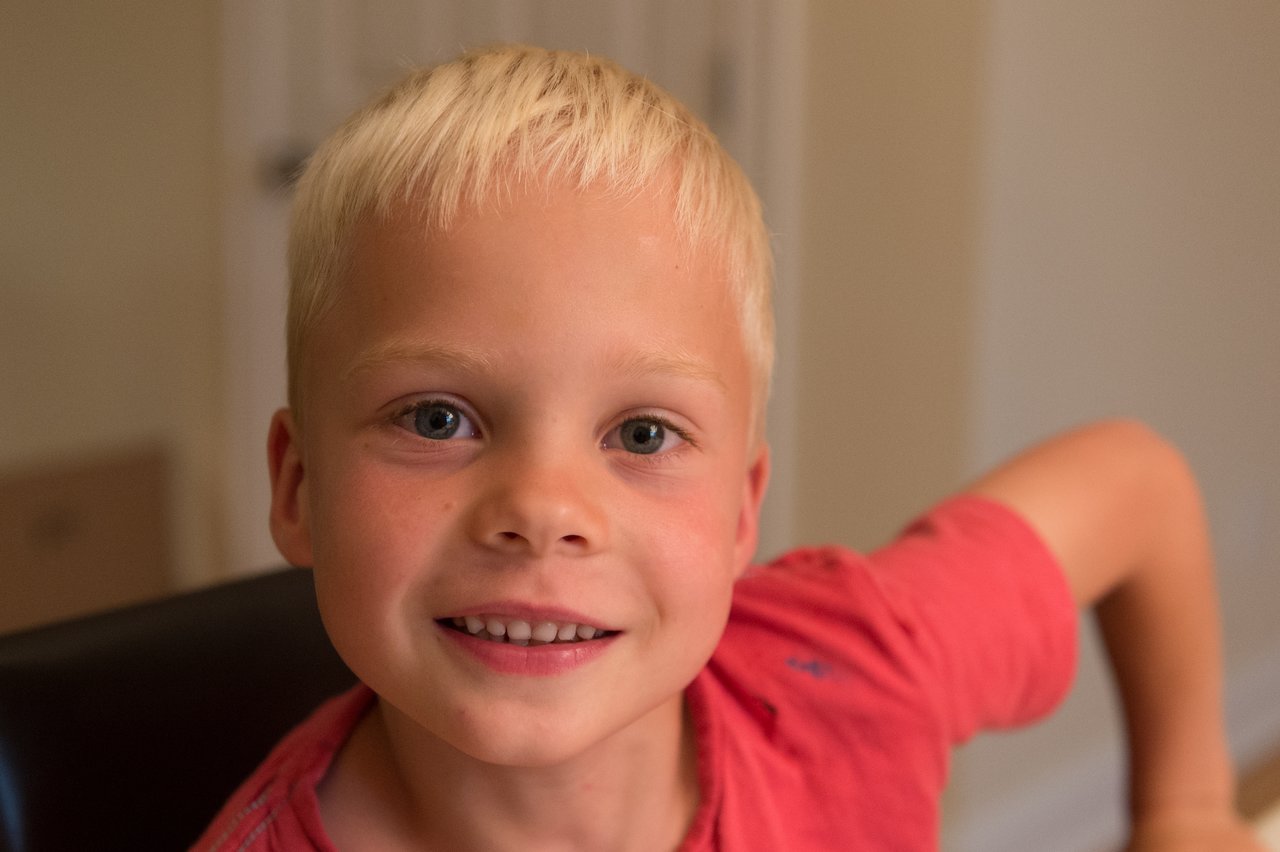 A smiling child in a red shirt sits indoors, looking at the camera during a birthday celebration.