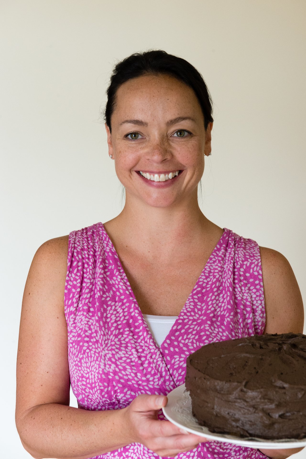 A smiling woman holds a chocolate cake on a plate, standing against a plain background.