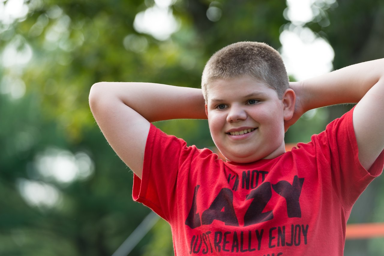 A boy in a red shirt smiles with his hands behind his head at an outdoor birthday party.