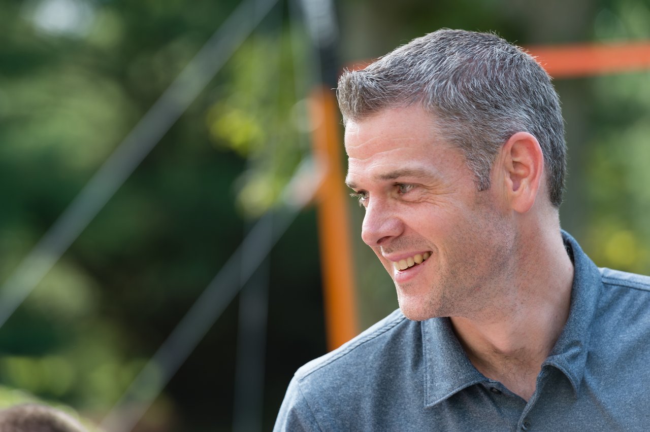 A man in a gray polo shirt smiles while looking to the side at an outdoor birthday party.