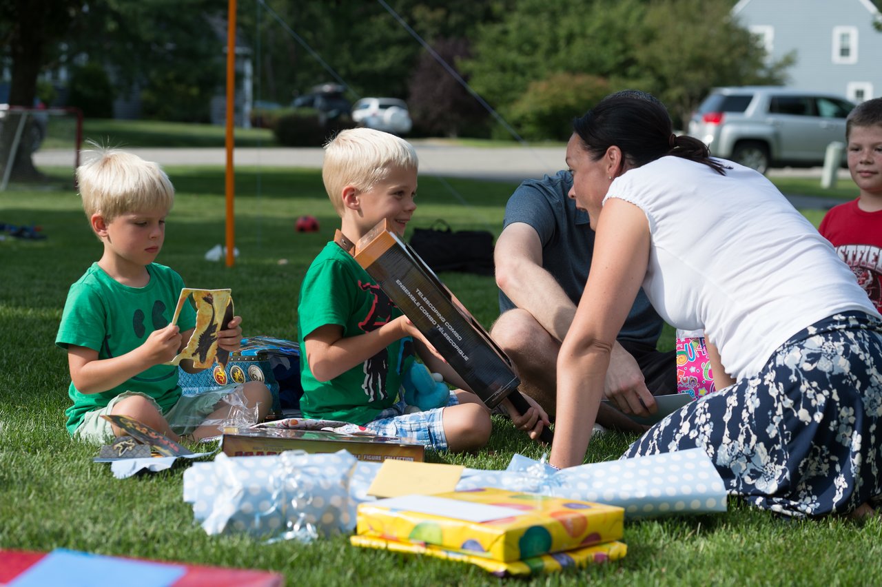 Two young boys sit on the grass opening birthday presents, while a woman and others watch and interact.