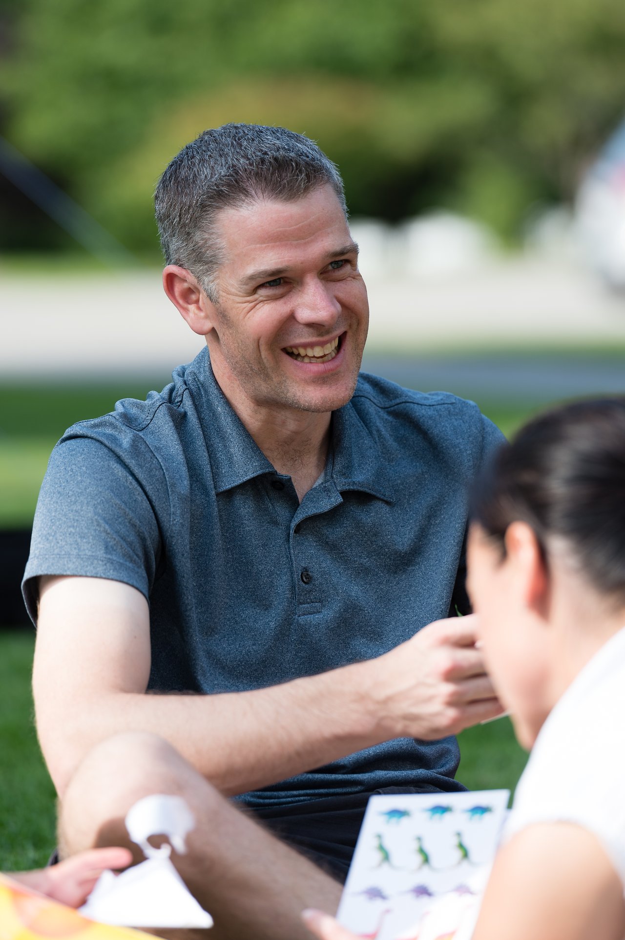 A man in a gray shirt smiles while talking to someone holding a sheet of stickers at an outdoor gathering.