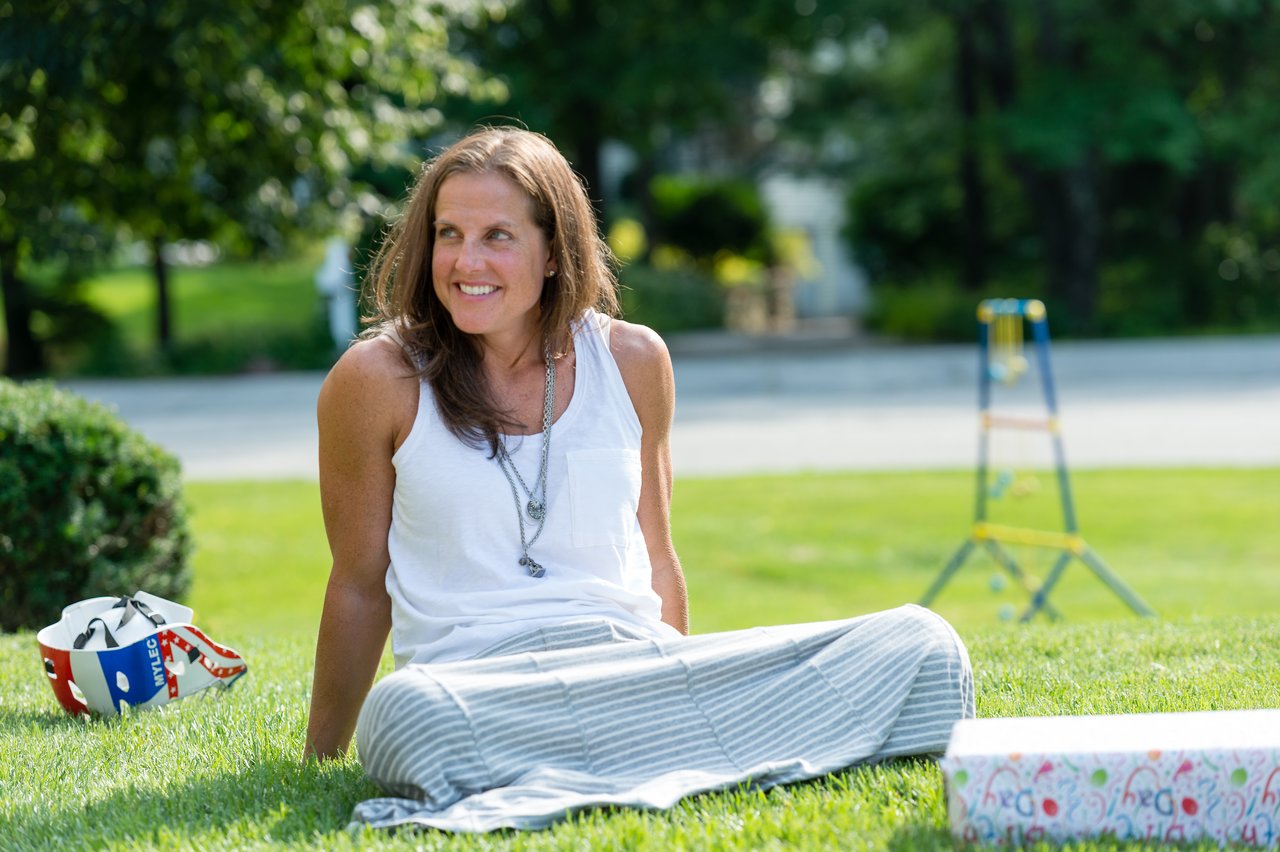 A woman sits on the grass, smiling, with a gift box nearby at an outdoor birthday party.