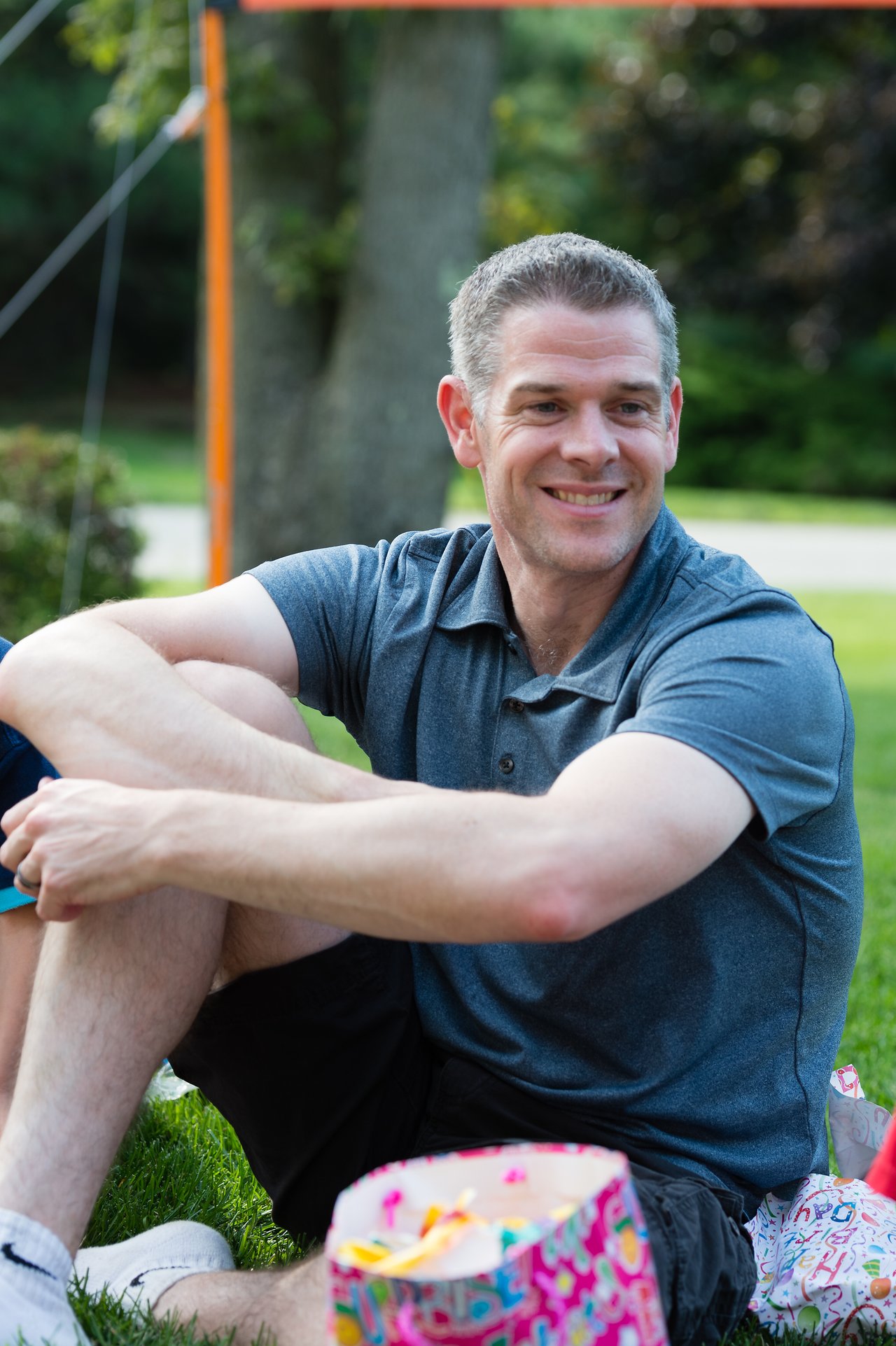 A man sits on the grass, smiling, with a colorful gift bag in front of him at an outdoor party.