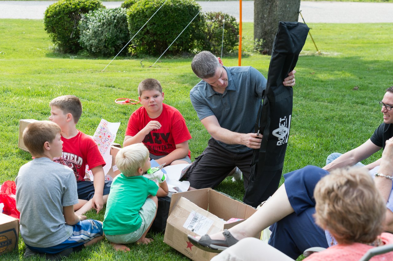 A group of children and adults sit on the grass while a man opens a black Rocket-branded bag.