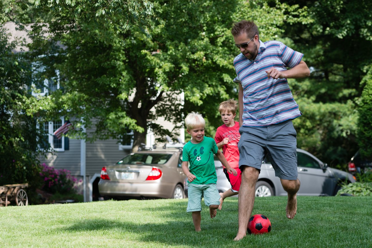 A man and two young boys play soccer barefoot on a grassy lawn during a birthday party.