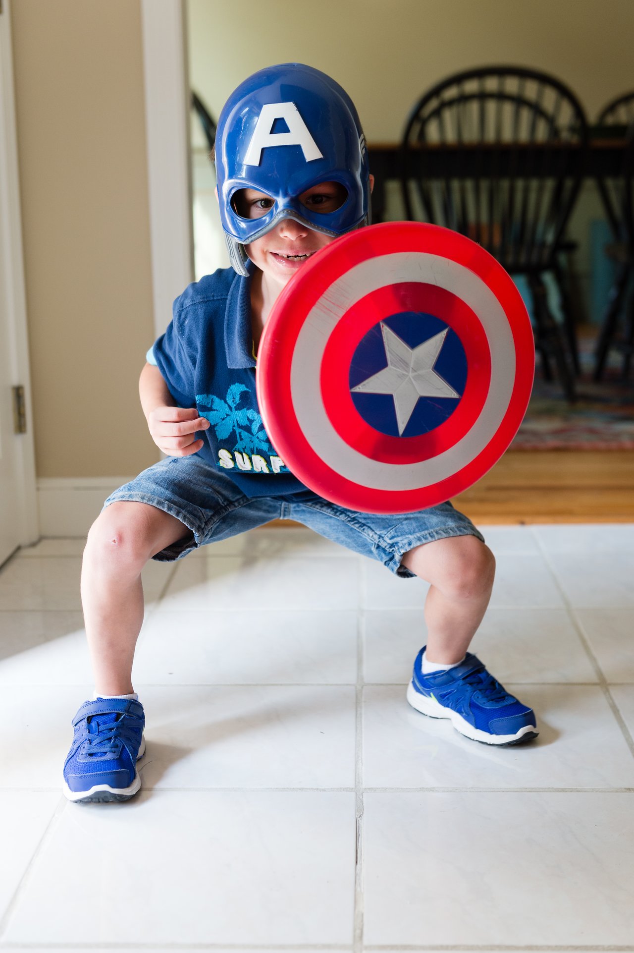A child wearing a Captain America mask holds a matching shield and poses playfully indoors.