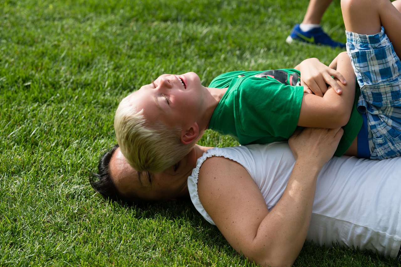 A child in a green shirt laughs while playfully lying on top of an adult on the grass.