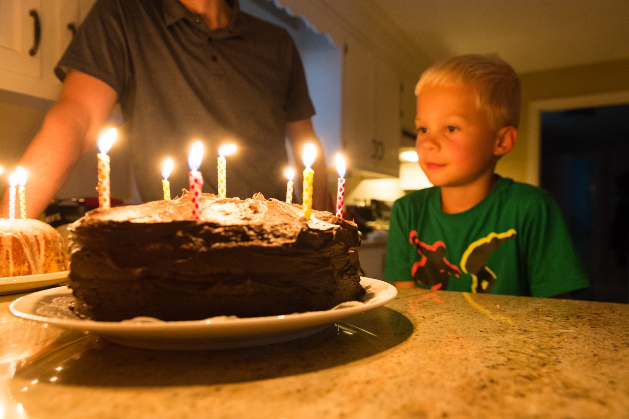 A young boy in a green shirt looks at a chocolate birthday cake with lit candles on a kitchen counter.