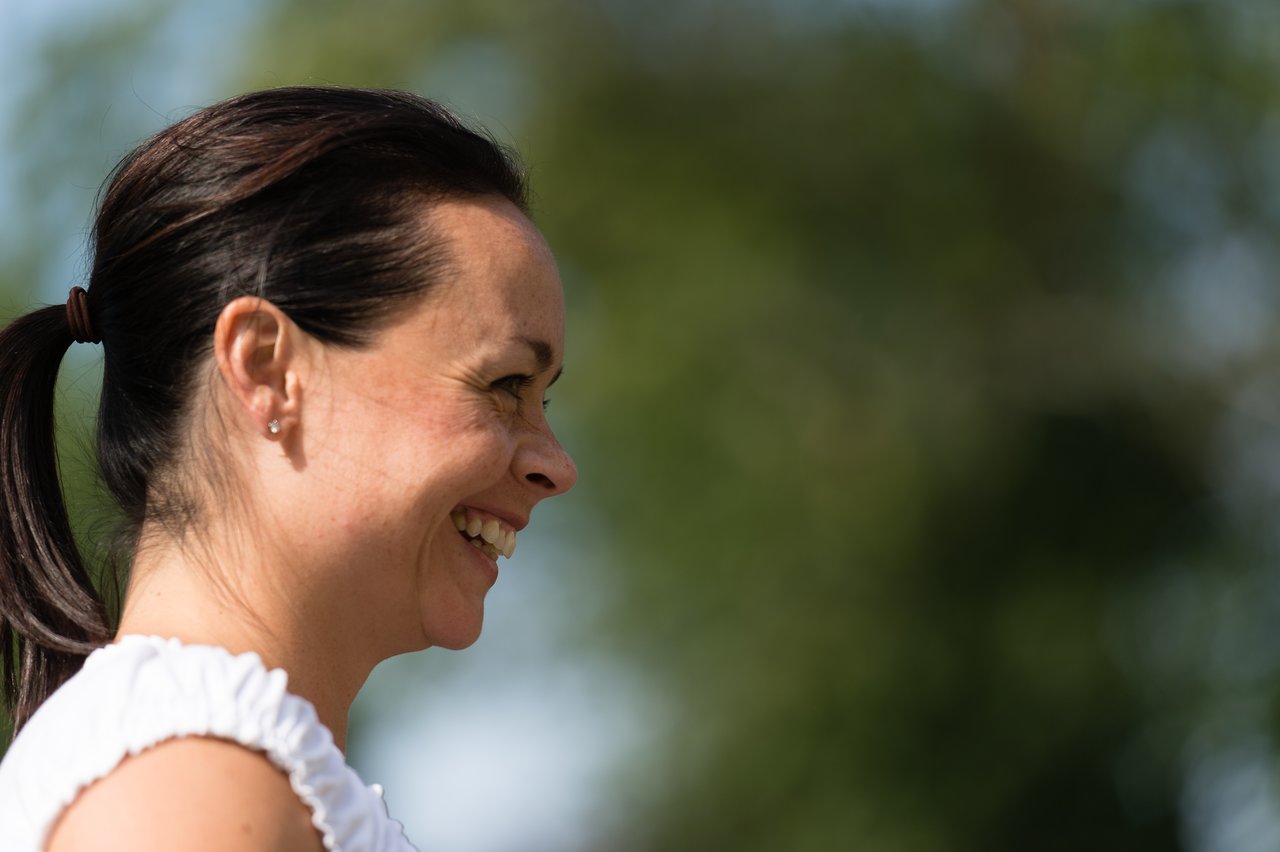 A woman with dark hair in a ponytail smiles while looking to the side at an outdoor gathering.
