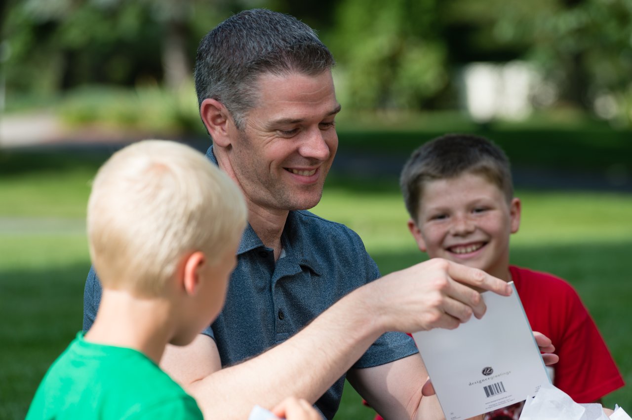 A man and two boys smile while opening a birthday card at an outdoor party.