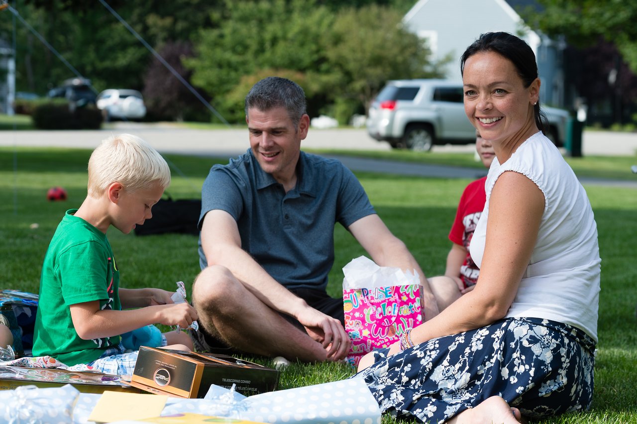 A young boy opens presents on the grass while two adults and another child smile and watch.