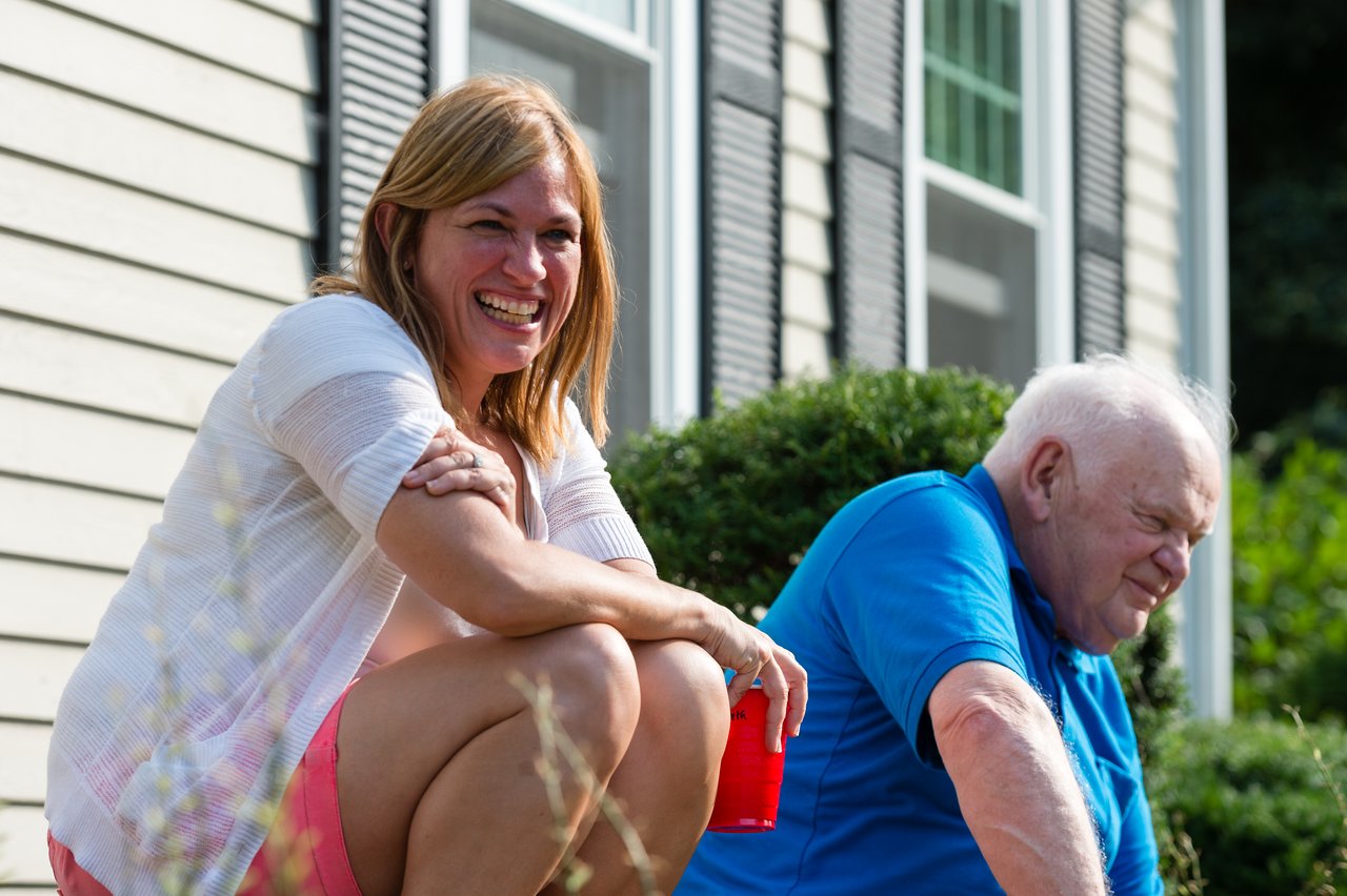 A woman and an older man sit outside a house, smiling and talking.
