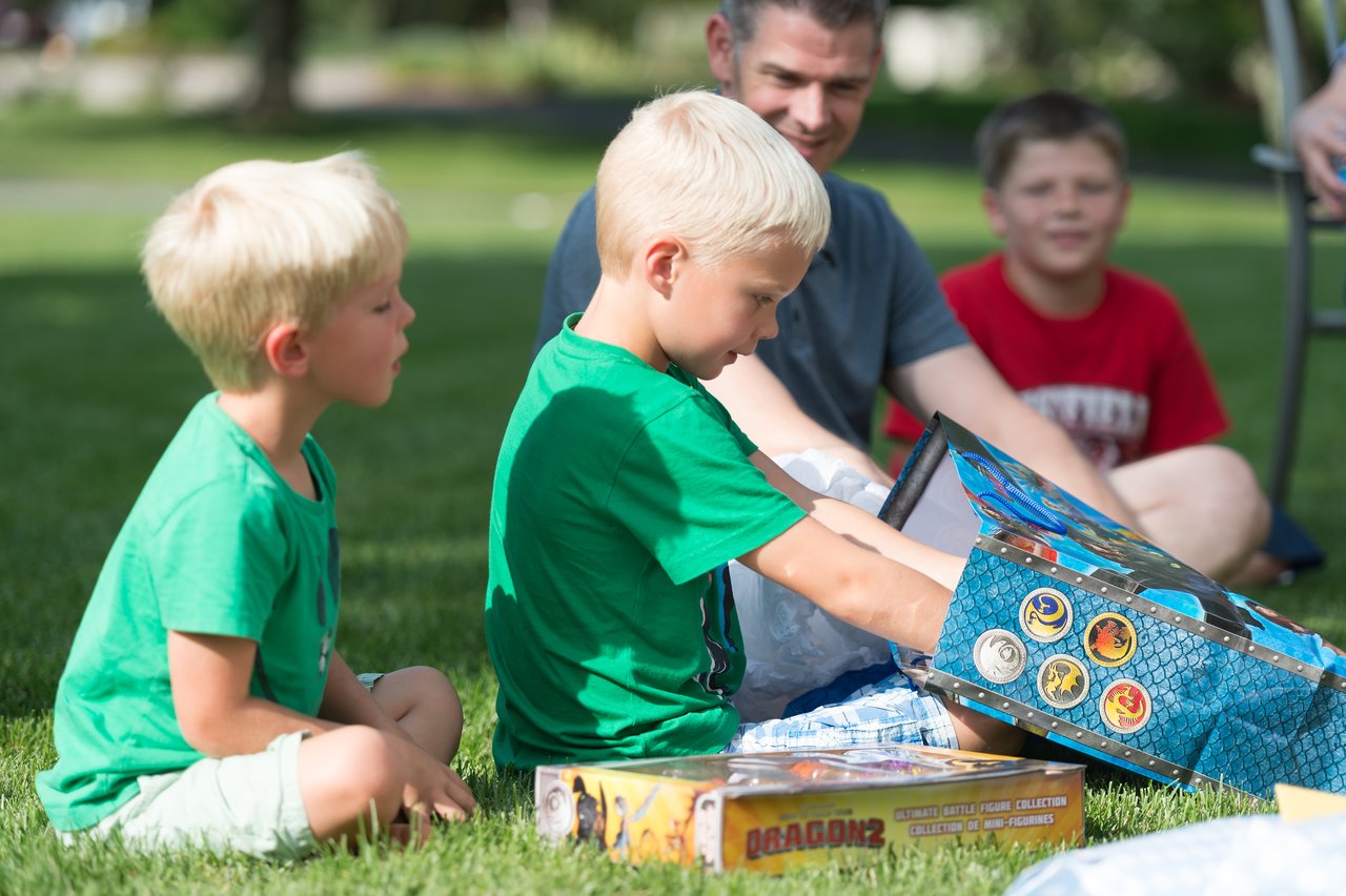 A young boy opens a gift box at a birthday party while another child and two adults watch nearby.