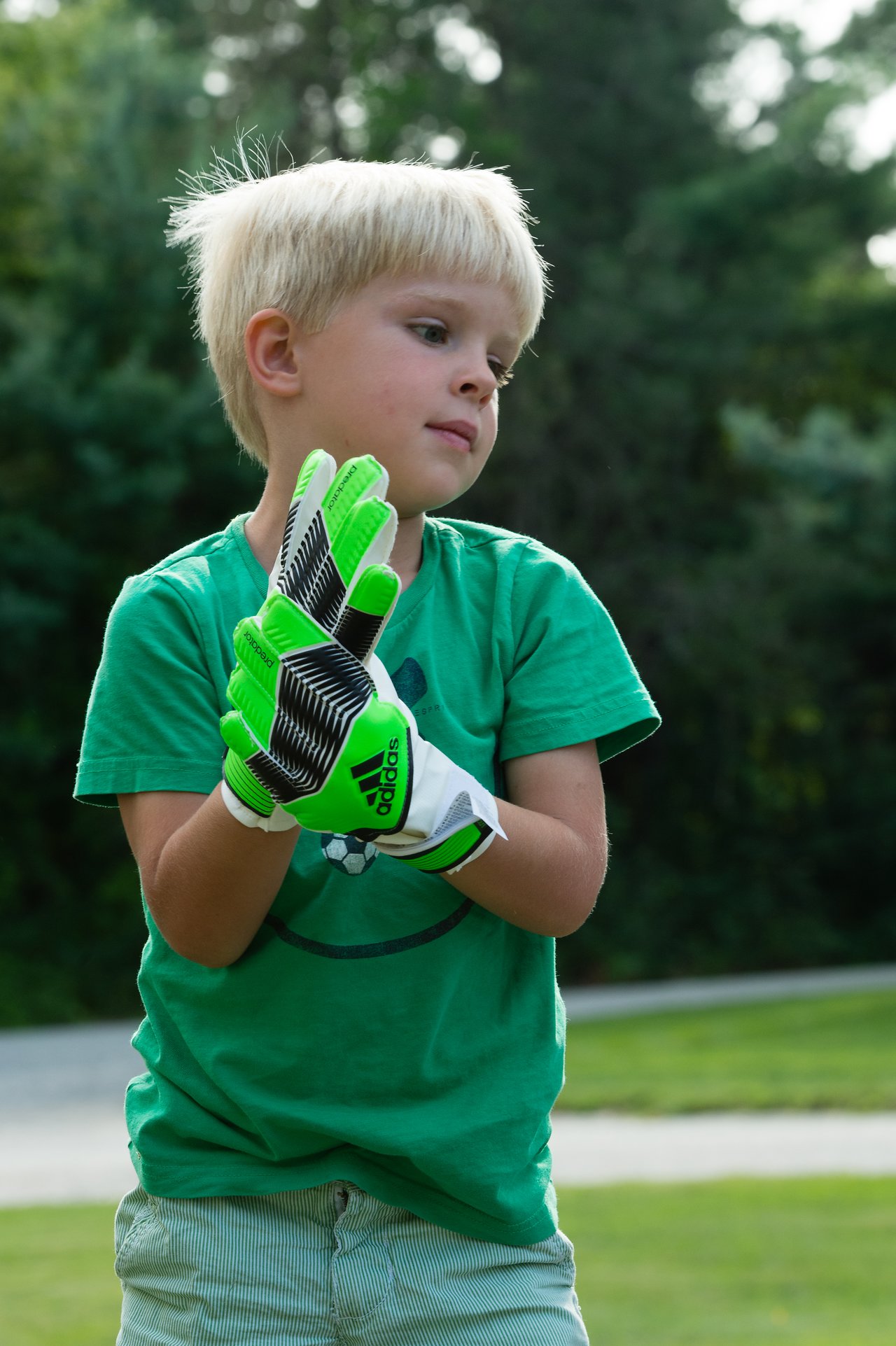 A young child wearing green goalkeeper gloves claps hands together while standing outside in a grassy area.