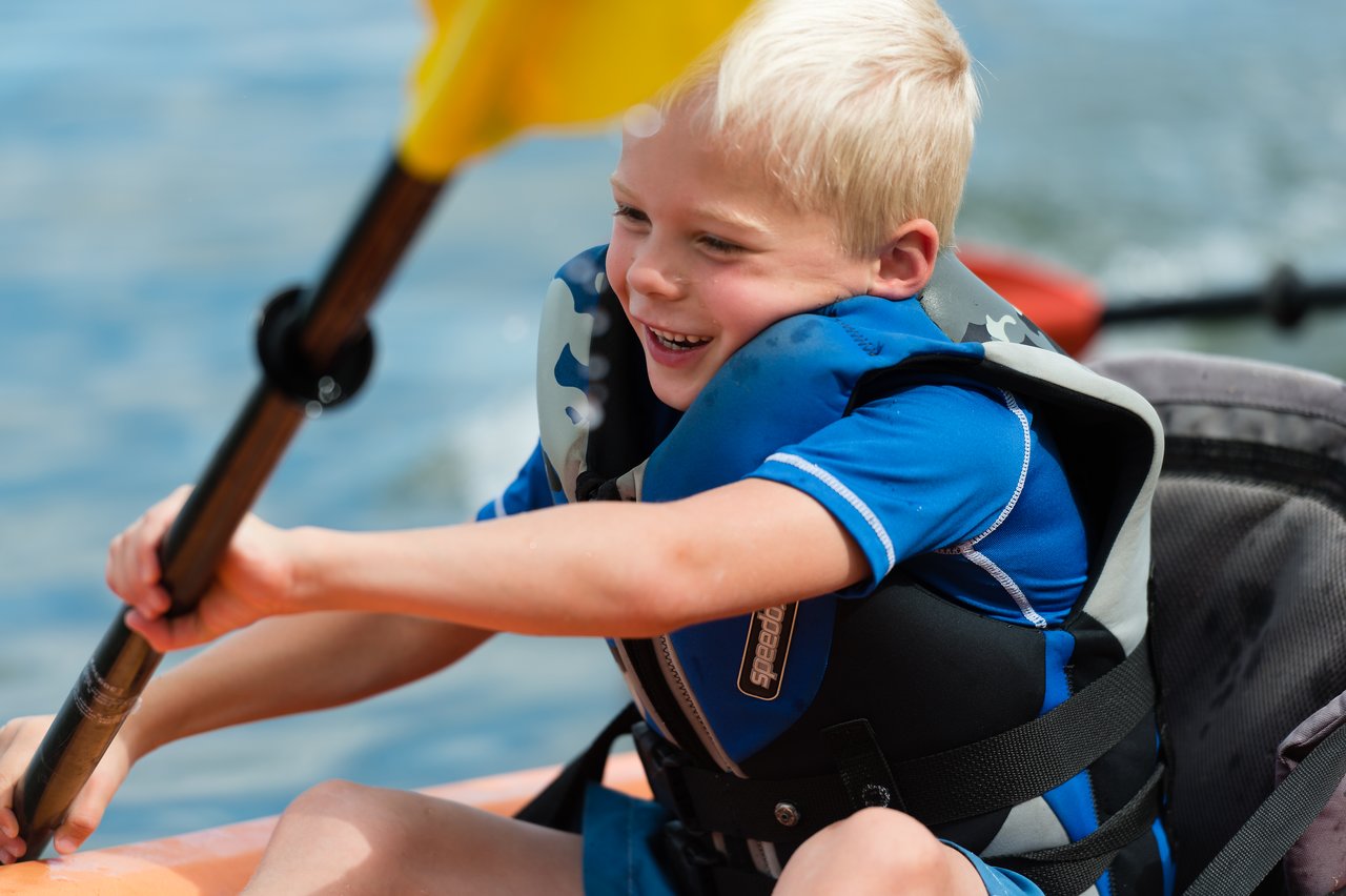 A young child wearing a life jacket smiles while paddling a kayak on the water.