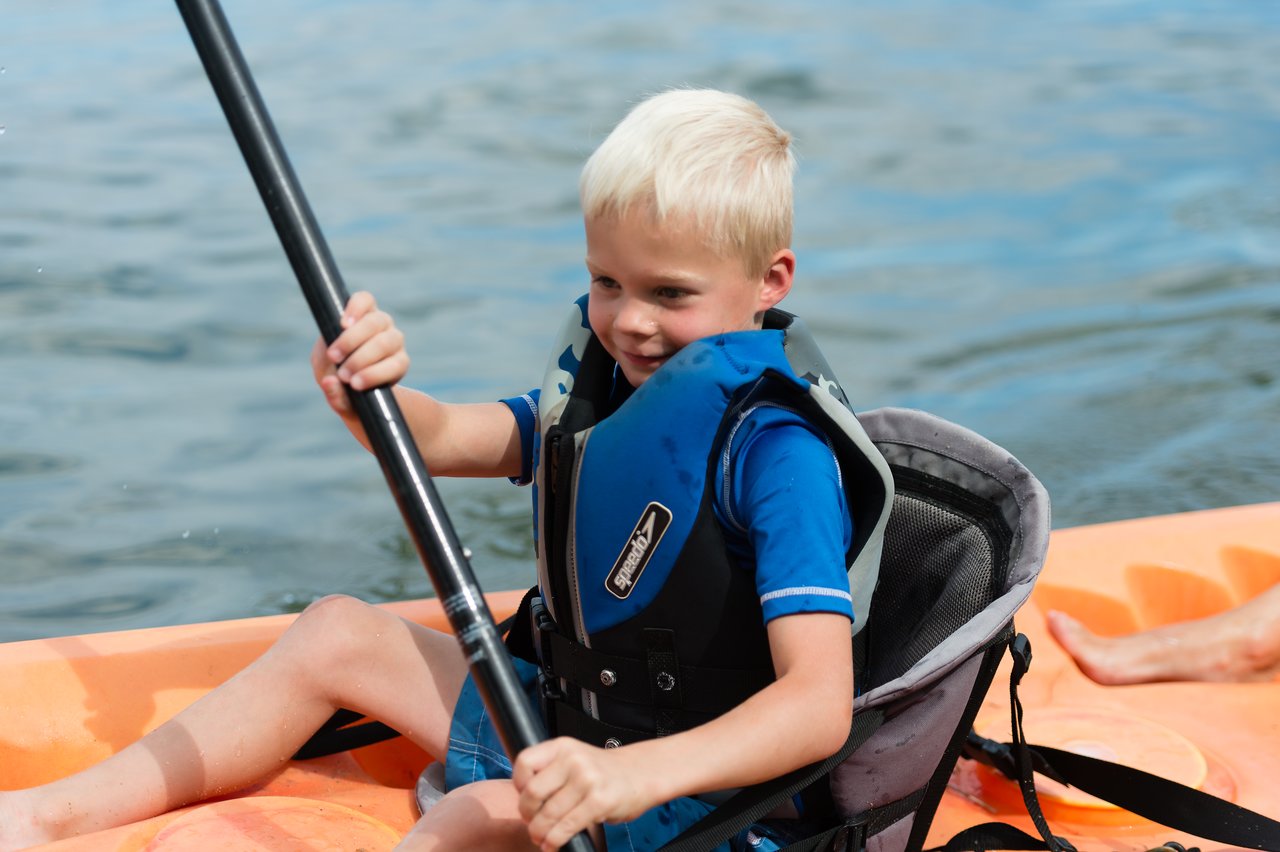 A young child wearing a life jacket paddles an orange kayak on the water, smiling and focused.